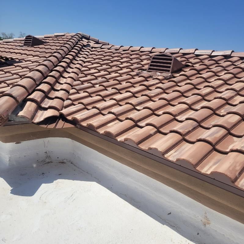 A close-up view shows a red Mediterranean tile roof section intersecting with a lighter flat roof area under a clear blue sky.