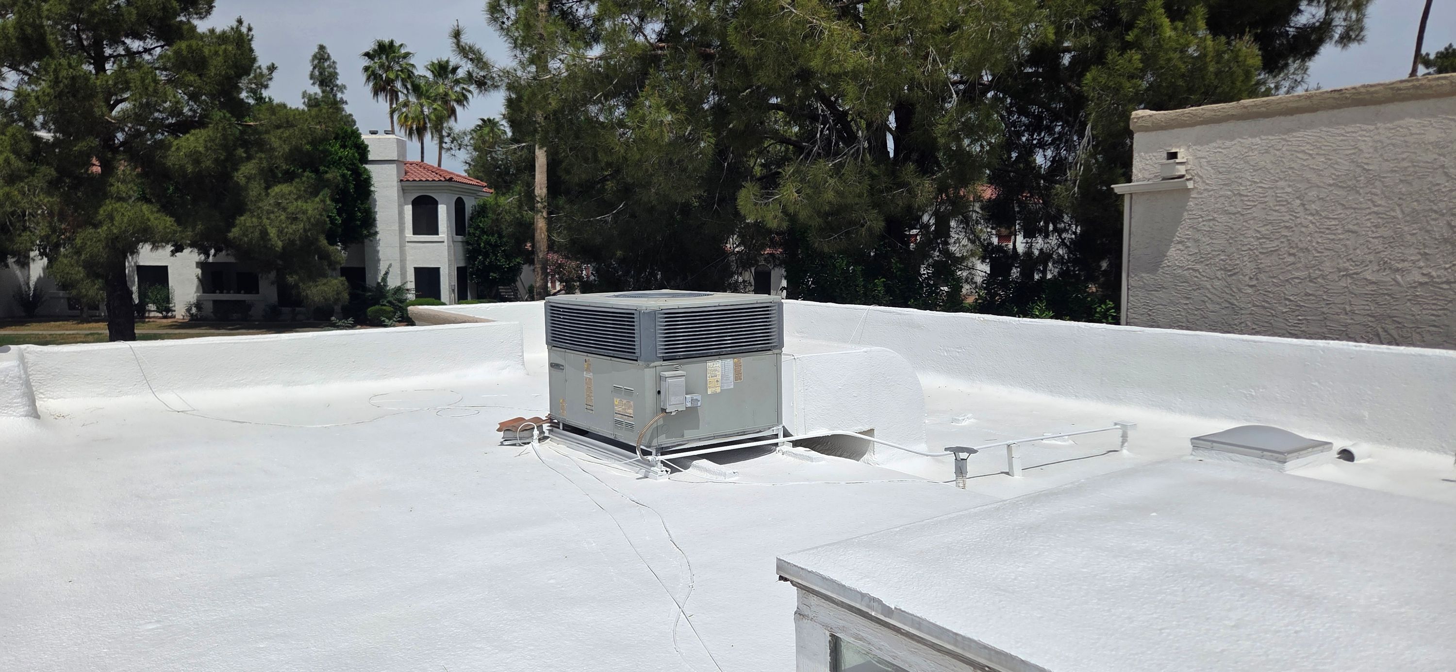 A newly installed white foam roof with an HVAC unit sits on a commercial building in Phoenix, surrounded by green trees.